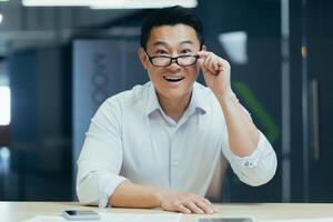 Portrait of a happy young Asian man sitting in the office at the table with phone and documents, holding glasses in his hand, looking down, smiling, looking at the camera. photo