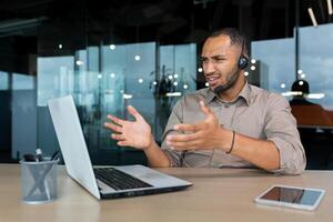 Angry worker with call headset talking to customer, hispanic man explaining angry and upset, businessman inside modern office with laptop for customer service support tech. photo