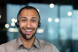 Close up hispanic male worker with headset smiling and looking at camera, tech support worker working inside office. photo