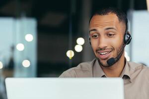 Close-up photo. Learning online. A young handsome African-American student is studying remotely from a laptop, sitting at a desk in a headset, talking on a call. photo