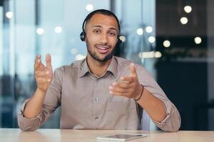 Webcam view, happy african american man talking online call looking at camera using headset, man working inside modern office building tech support and helpline call center. photo