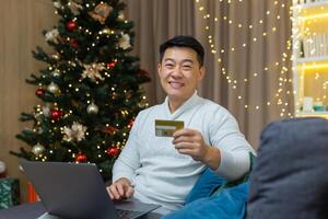 Man with bank credit card smiling and looking at camera with laptop sitting on sofa in living room at home near Christmas tree, portrait of happy asian shopper in online store for new year. photo