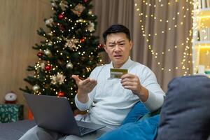 Upset christmas man with laptop trying to make online purchase in online store, asian man holding bank card received error and fraud message, sitting on couch for new year. photo