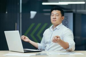 Portrait of Asian businessman, man in office working with laptop, meditating at workplace with eyes closed photo