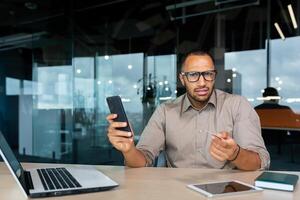 Dissatisfied businessman inside office looking at camera, hispanic man holding bank credit card and phone in hands, cheated man got rejection and error when trying to make online money transfer. photo