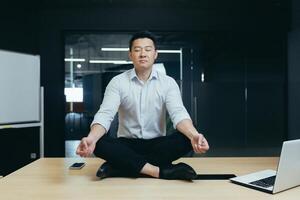 Asian boss businessman meditating in office, man sitting in lotus position on desk with closed eyes, thinking about important decision photo