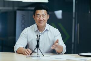 Asian smiling young man sitting in office at desk in front of camera, talking on microphone via call, blogging, webinar, training, recording podcast. photo