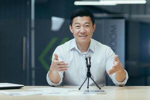Close-up photo portrait of businessman man looking into web camera and smiling using professional microphone for communication and online podcast recording, Asian business coach