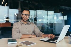 Portrait of successful businessman, man with laptop working inside modern office building, startup entrepreneur working with laptop, wearing glasses smiling and looking at camera. photo