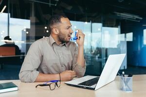 Sick office worker with asthma having trouble breathing, man using inhaler to relieve breathing, hispanic man working inside modern office building, mixed race man with laptop photo