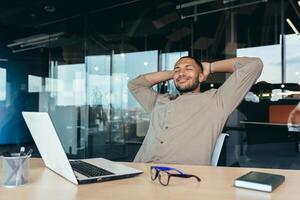 mixed race man relaxing in office with hands behind head looking out window and smiling, businessman daydreaming inside modern office building, successful investor happy with work. photo