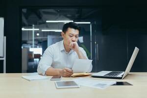 Stressed young asian businessman working from office report annoyed with low sales. Nervous boss see c mistake incorrect information in paper document photo