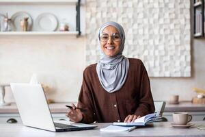 Portrait of a young Muslim female psychologist specialist in a hijab sitting at home in front of a laptop and making notes in a notebook. He consults and works online. Smiling looking at the camera. photo