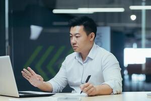 Worried Asian businessman sitting at the table in the office with laptop, talking on a call online, holding a pen, working with documents. solving the problem with the bank, bills, credit. photo