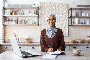 Portrait of a young female student of oriental origin in a hijab sitting at home in the kitchen and studying remotely from a laptop. Notes lectures, lessons. Smiling, she looks at the camera. photo