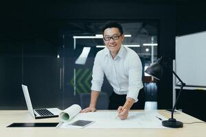 Portrait of successful and experienced Asian architect, man standing over table with plan drawing, smiling and looking at camera, working in modern design studio photo
