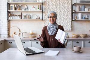 Distance Learning. A young Arab woman in a hijab is teaching online at home via a call from a laptop. Shows the camera a book, explains, reads a lecture. He looks at the camera with a smile. photo