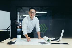 Portrait of a young Asian man architect, designer, engineer standing in the office at the desk, working with documents, projects. He looks at the camera with a smile. photo