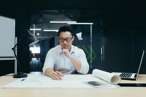A handsome young man, an Asian architect, designer, engineer, sits at a desk in the office at a table with notebooks and documents. He concentrates on making drawings, developing a plan. photo