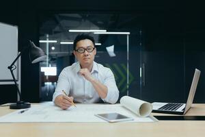 Portrait of a young Asian male designer, architect, engineer looking confidently and seriously at the camera, working in the office at a table with papers and projects. photo