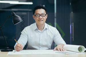 Portrait of young Asian man wearing glasses, designer, architect, looking at camera, smiling. Sitting at a table in a modern office, working with drawings, holding a pencil in his hands. photo