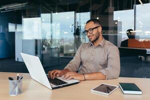 African american programmer at work with laptop inside office, man in glasses and casual writing code for programs. photo