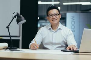 Portrait of Asian designer, creative work, man looking at camera and smiling, working in architecture office on plan drawing photo