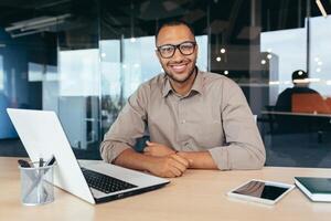 Portrait of successful investor, african american man smiling and looking at camera, man working inside office using laptop at work, satisfied with achievement result. photo