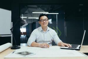 Portrait of successful asian male architect smiling and looking at camera, working on project plan drawing in design studio office photo