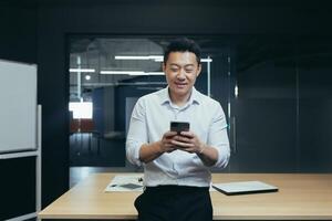 A smiling young Asian businessman is standing in the office at a desk and using a mobile phone, writing a message, searching for information on the Internet, checking social networks. photo