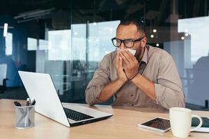 Man sneezes and has runny nose in office at work african american businessman sick working inside office at desk using laptop. photo
