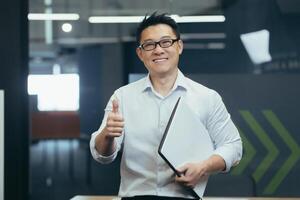 Portrait of a successful young Asian male programmer standing in a modern office with a notebook in his hands, smilingly looking at the camera, showing the super finger. photo