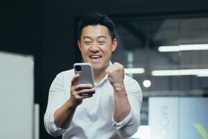 A break at work. A young Asian man holds a phone in his hands, plays games on the phone in the office at work, rejoices, shows with his hand yes,rejoices at the win, won a prize, rests, does not work. photo