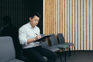 A young Asian man sits in the lobby and fills out a survey questionnaire in the medical center, in the office for work, interview, arrival at the hotel. photo