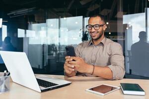 Happy and smiling businessman rejoicing at successful work results and achievement, man at work using phone reading online good news sitting at table inside office, using laptop at work. photo