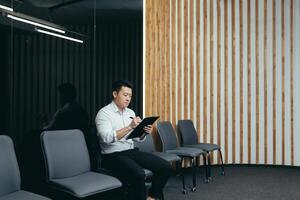 A young handsome Asian man is sitting seriously in the lobby of the office center, waiting for an interview, holding a folder with papers and documents in his hands. He is filling out a questionnaire. photo
