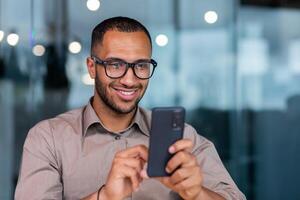 Close-up of smiling African-American businessman using phone typing messages and browsing internet pages, man in shirt and glasses smiling happily inside office holding smartphone. photo