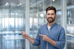 A smiling, bearded man points with both hands, suggesting something is to his side in an office setting. photo