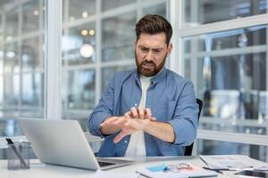 A man at his desk is visibly irritated, scratching his arm while working. The workspace includes a laptop, documents, and a window view. photo