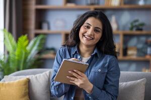 Close-up portrait of a young smiling Indian woman at home, looking at the camera and taking notes in a notebook. photo