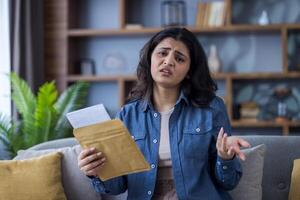 A young woman looks upset as she reads a document that she has just pulled from a brown envelope while at home on the couch. photo