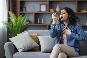 Young woman using an inhaler while sitting on a couch in her living room, experiencing an asthma attack and shortness of breath. photo