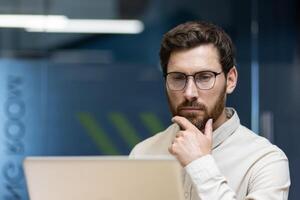 Close-up photo of a thoughtful young businessman sitting in the office at the workplace and looking at the laptop screen while rubbing his beard with his hand.