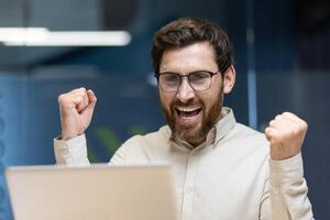 Close-up photo of a young happy man who is in the office, looking at the laptop screen and is happy, showing a victory gesture with his hands.