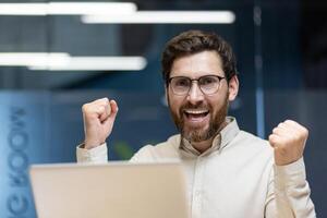 A close-up portrait of a young happy and successful businessman who is in the office, sitting at the workplace and is happy to the camera, showing a victory gesture with his hands. photo