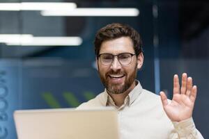 Close-up photo of a young smiling man sitting in an office at a workplace in front of a laptop, greeting and waving at the camera.