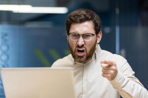 Close-up photo of a young angry man sitting in the office at the desk and shouting while pointing his finger at the laptop screen.