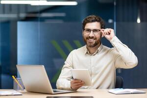 Portrait of a young smiling man working in the office, sitting at a desk with a laptop, holding glasses, using a tablet and looking at the camera. photo