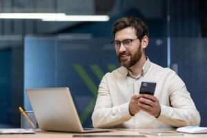 Smiling young man working in office, sitting at desk, using mobile phone and looking at laptop screen. photo