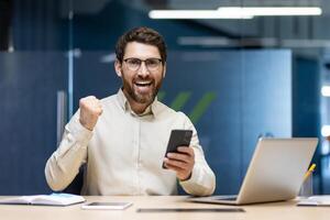Portrait of a happy young man businessman sitting in the office at the table with a laptop, using a mobile phone in his hands and rejoicing in happiness and achievement showing a yes gesture. photo